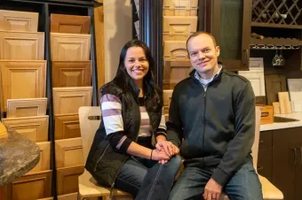 Two people sitting together in a showroom with stacked wooden cabinet doors, smiling at the camera.
