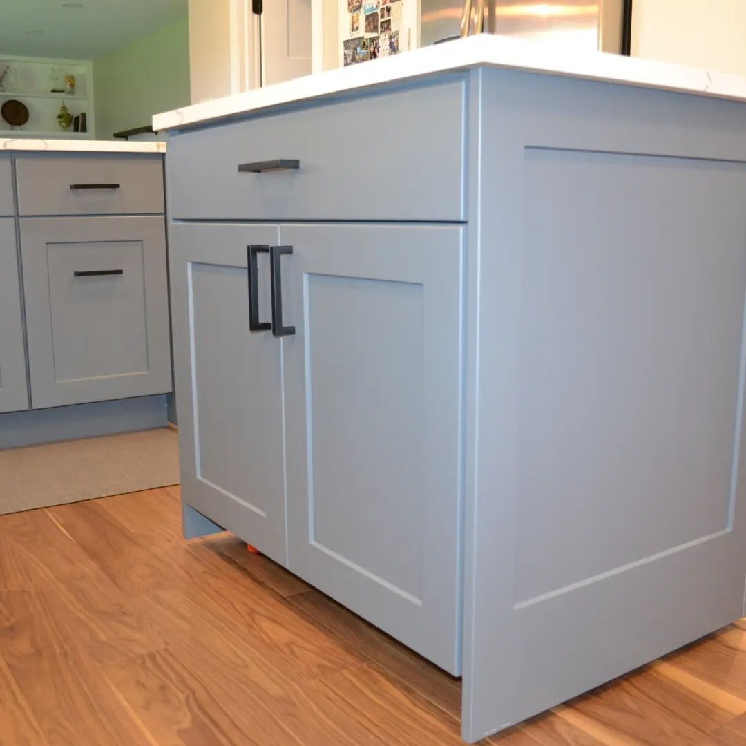 Gray kitchen cabinets with black hardware and a white countertop on a wooden floor.
