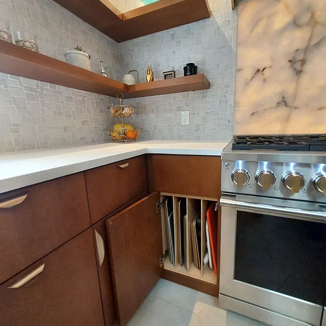 Kitchen corner with wooden cabinets, open oven, and baking sheets. Shelves and backsplash are light-colored.