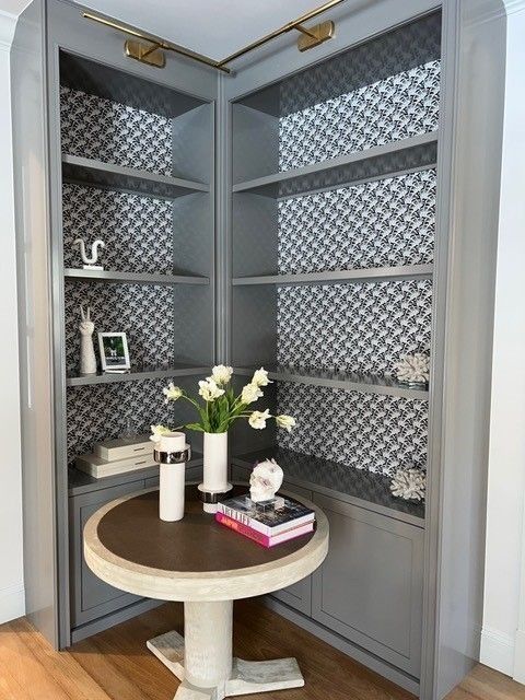 Corner bookshelf with a round table. Gray shelves with patterned backing, flowers and decor.