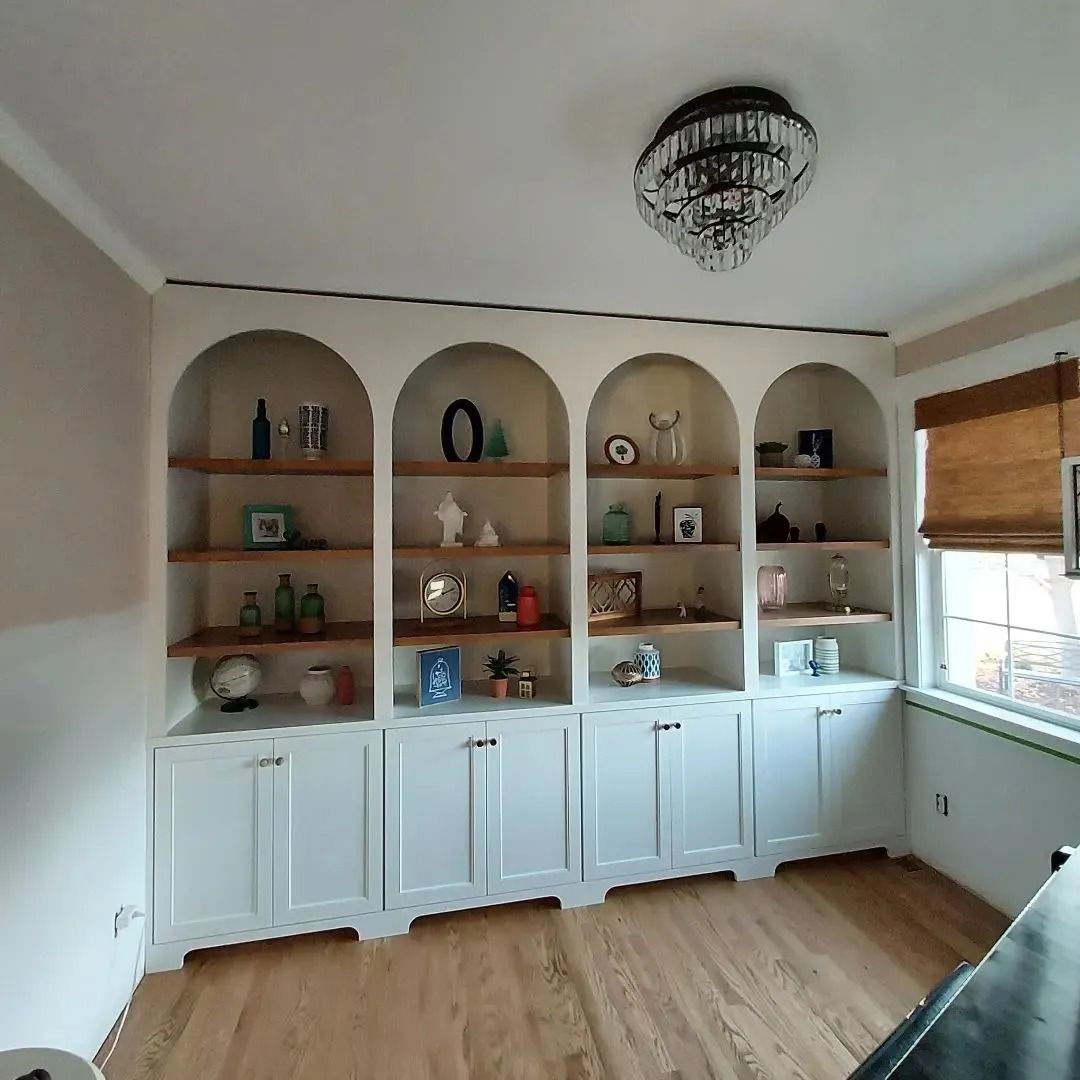 Built-in white bookshelves with arched openings, wooden shelves, and lower cabinets. A chandelier hangs above the shelves.