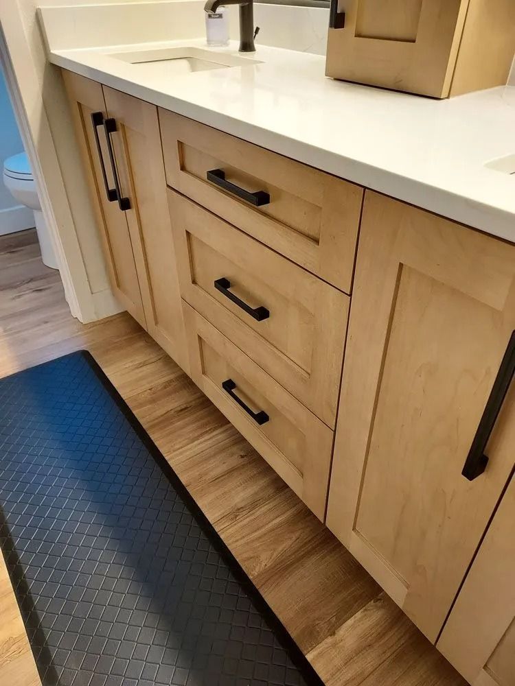 Bathroom vanity with light wood cabinets, black hardware, and a dark blue anti-fatigue mat.