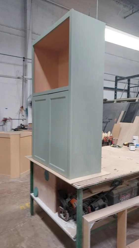 Tall, sage green cabinet with two doors and open top, sitting on a work table in a workshop.