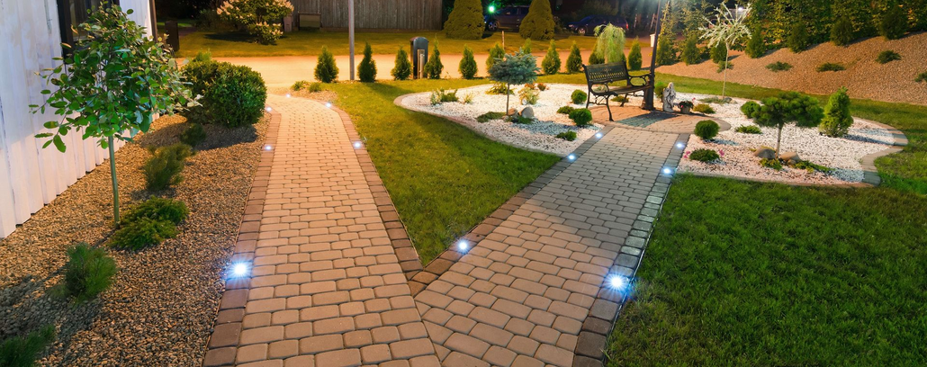Brick pathway lit by ground lights through a landscaped garden at night, leading to a patio and lawn