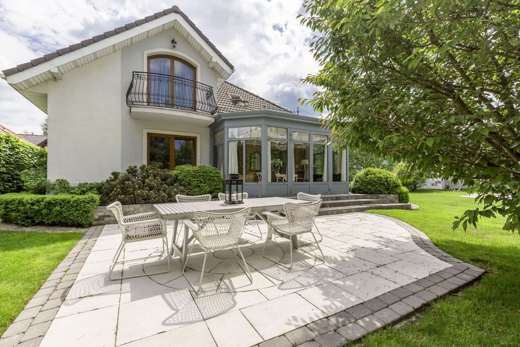 Patio with white metal table and chairs outside a white house with glass sunroom and green lawn