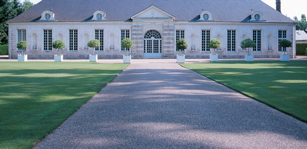Large stone mansion with a central arched entrance and a long gravel driveway, flanked by green lawns.