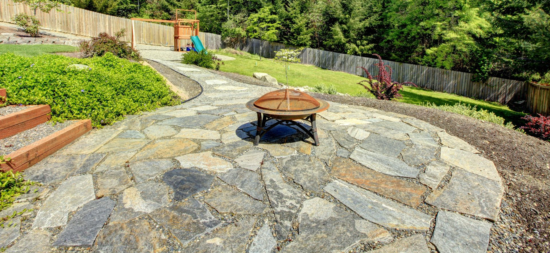 Stone patio with a central fire pit, curved path, and lush garden beds on a sunny day