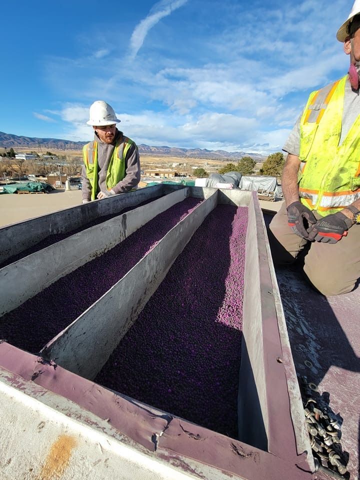 Two workers in safety gear inspecting purple gravel-filled troughs on a rooftop, sunny day.