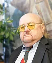 Bald man in a suit and yellow glasses, standing indoors with a blurred mural background