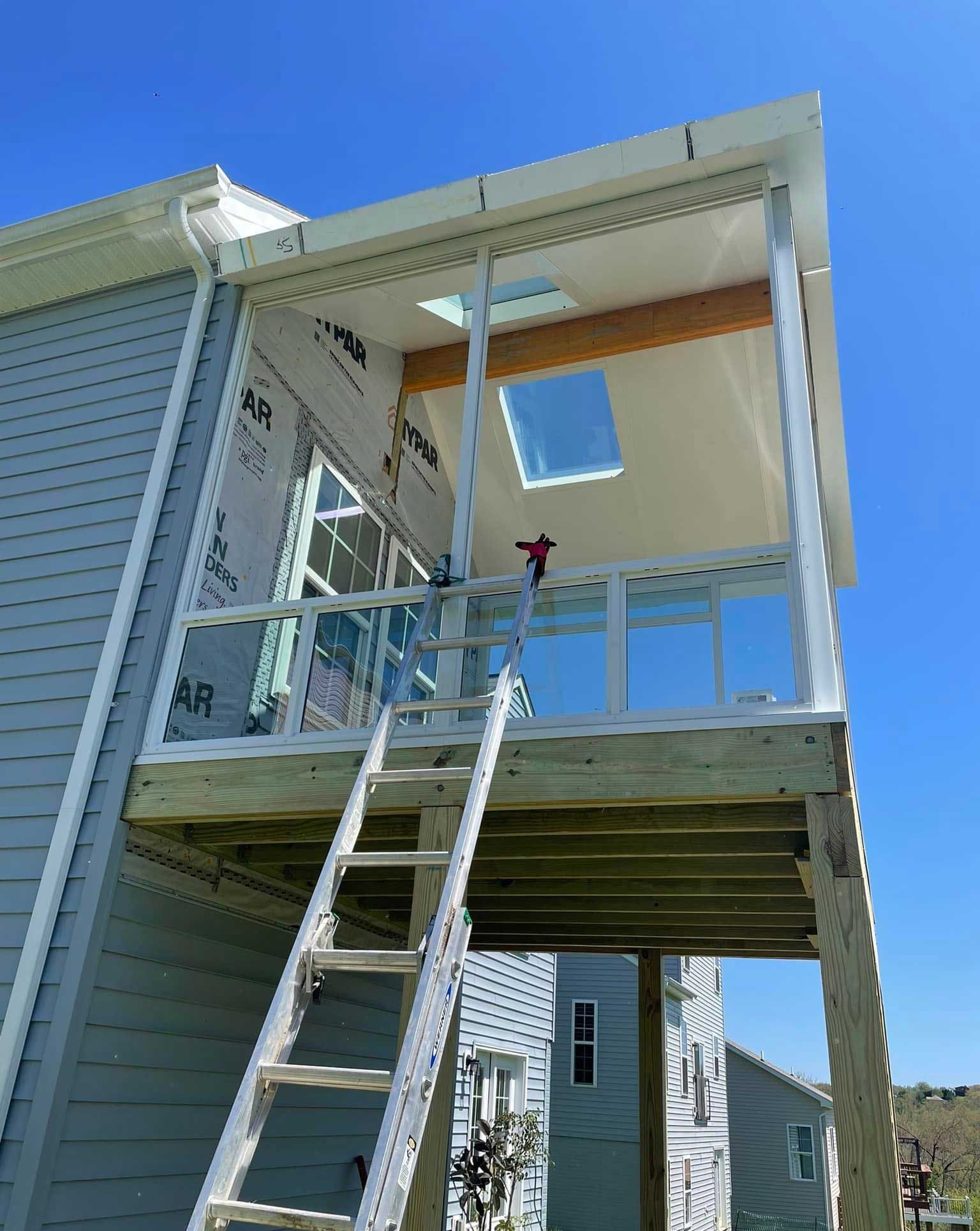 Ladder leads to a screened-in porch under construction attached to a blue-sided house, against a bright blue sky.