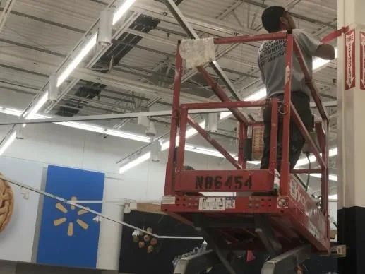 Worker on a red lift inspecting the ceiling inside a store, with a Walmart flag in view