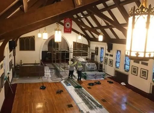 Gymnasium with high wooden ceiling, workers cleaning a polished floor and hanging lights.