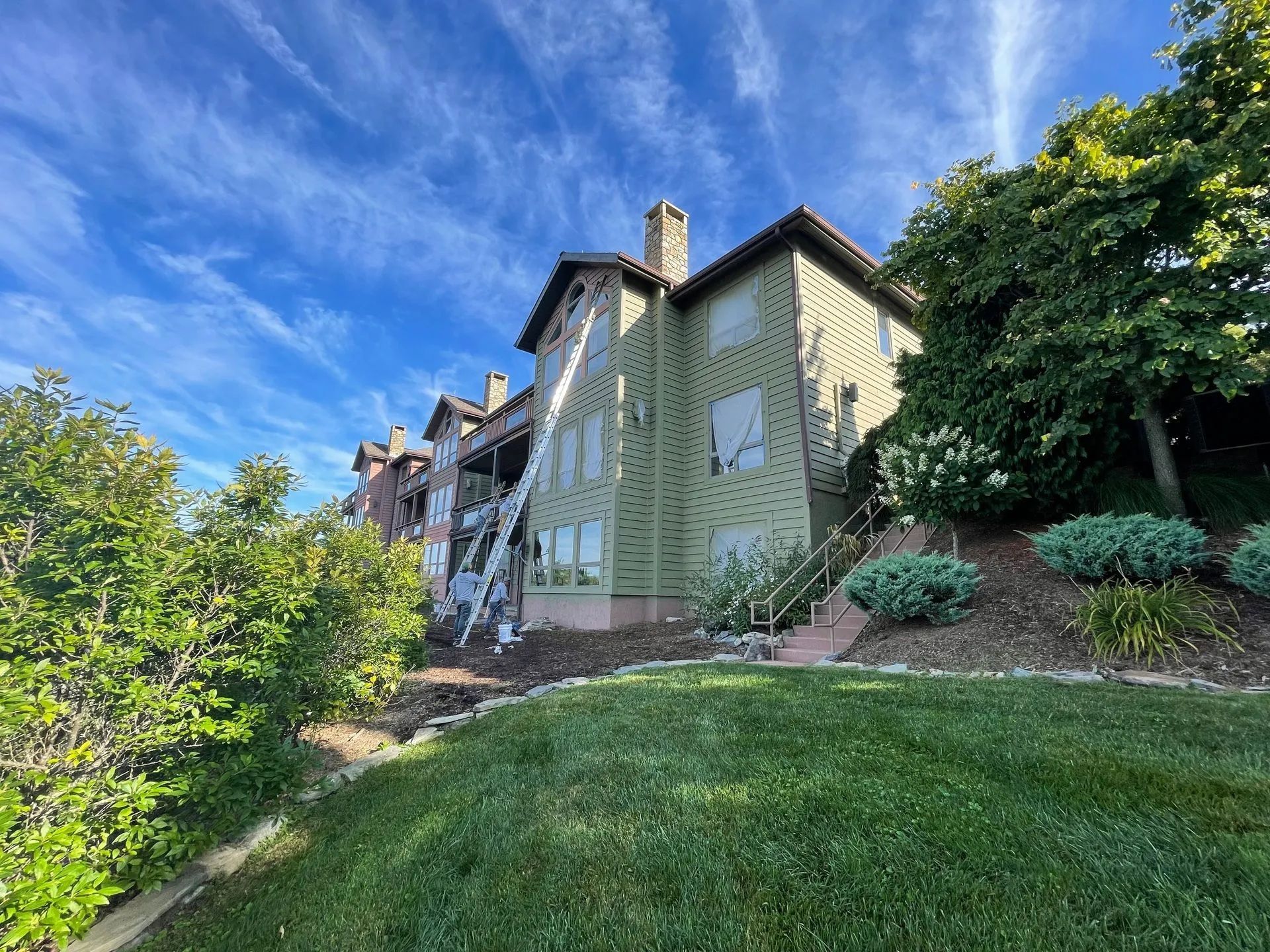 Large ivy-covered house on a hillside with a green lawn, trees, and a blue sky with wispy clouds.