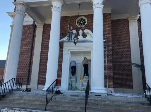 People installing a chandelier at the entrance of a brick building with white columns and steps.