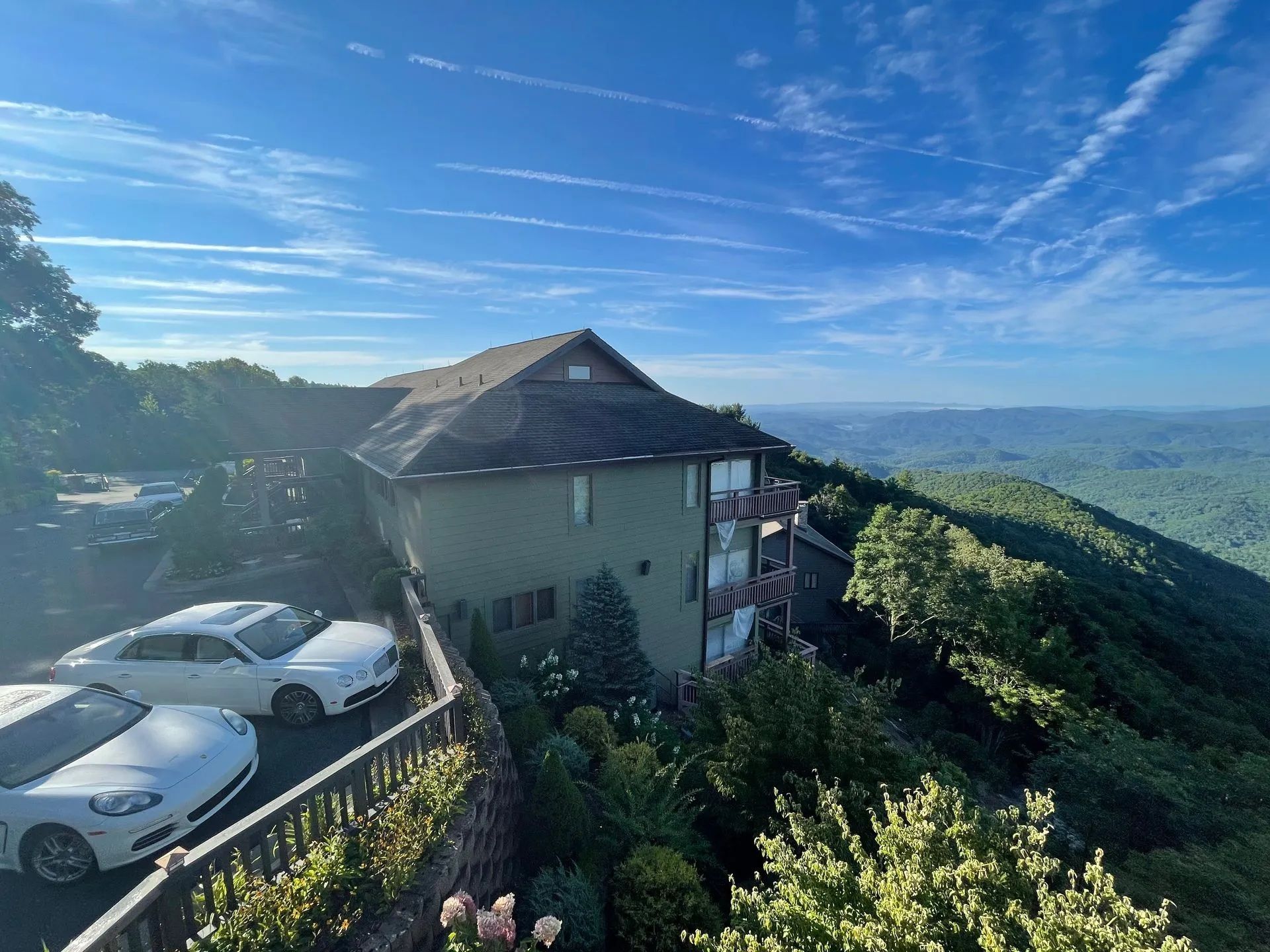 House on a hillside with parked cars, overlooking a lush valley under a blue sky