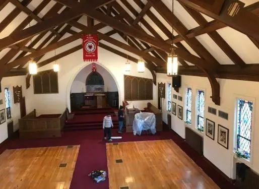 Church interior with wooden beams, altar, stained glass windows, and two people near the front.