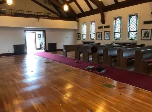 Empty church sanctuary with wooden floor, pews, stained-glass windows, and a maroon aisle runner