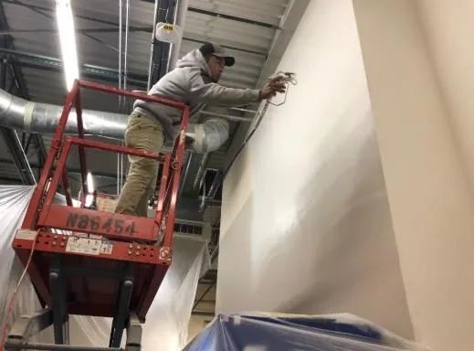 Worker in a red lift painting a tall white wall indoors