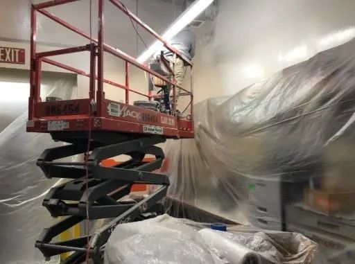 Worker on red scissor lift in a plastic-covered indoor space near a wall and exit sign.