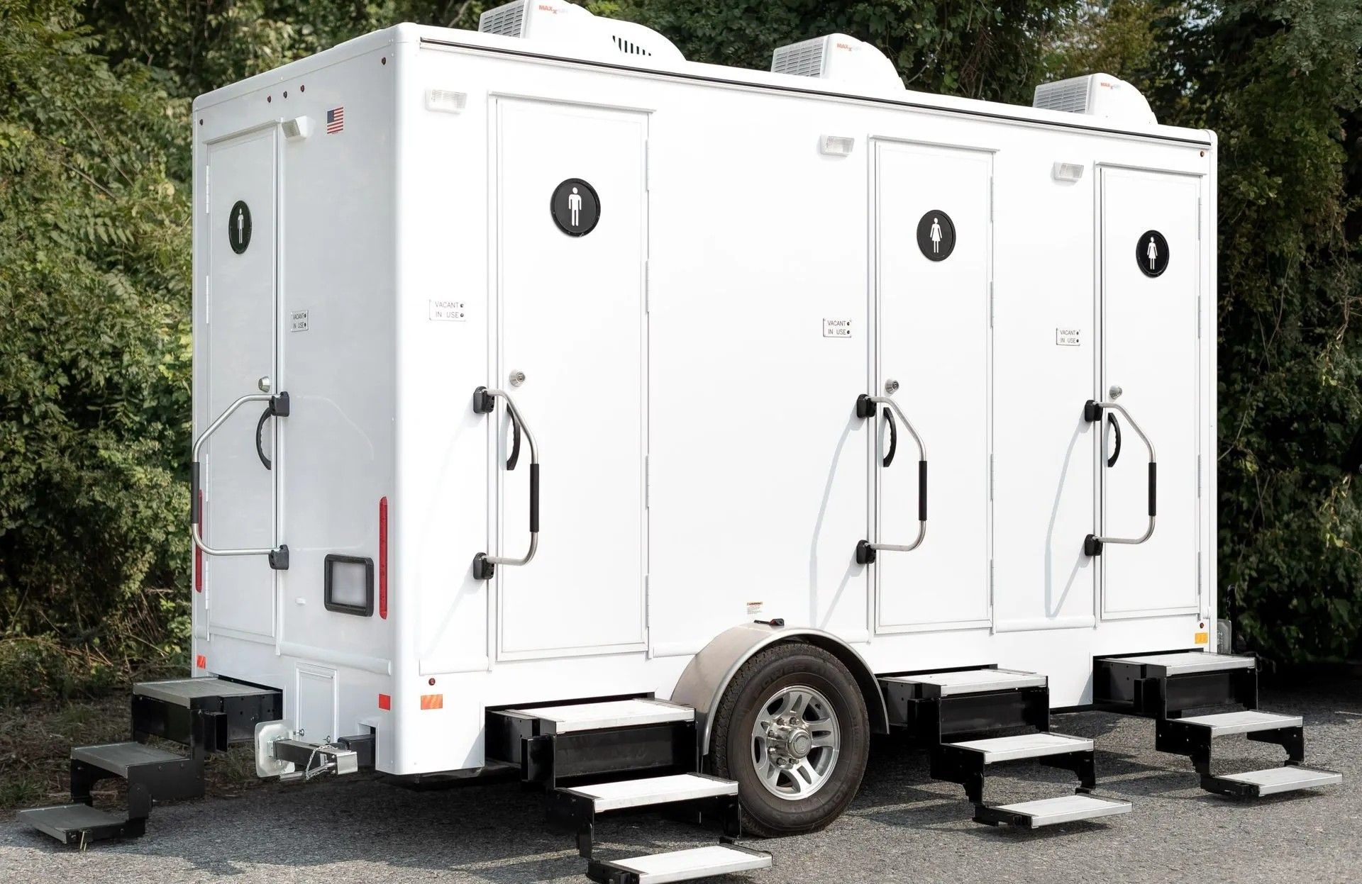 White portable restroom trailer with three stalls, steps, and black door symbols, outdoors.