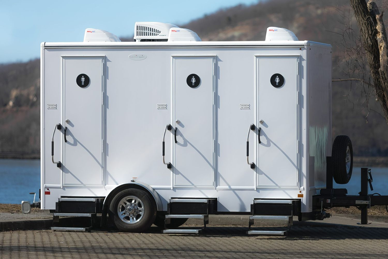 A white trailer with three toilets on it is parked next to a body of water