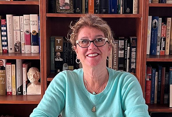 Woman with glasses smiles in front of a bookcase. She wears a turquoise sweater, jewelry. Bookshelves are brown.