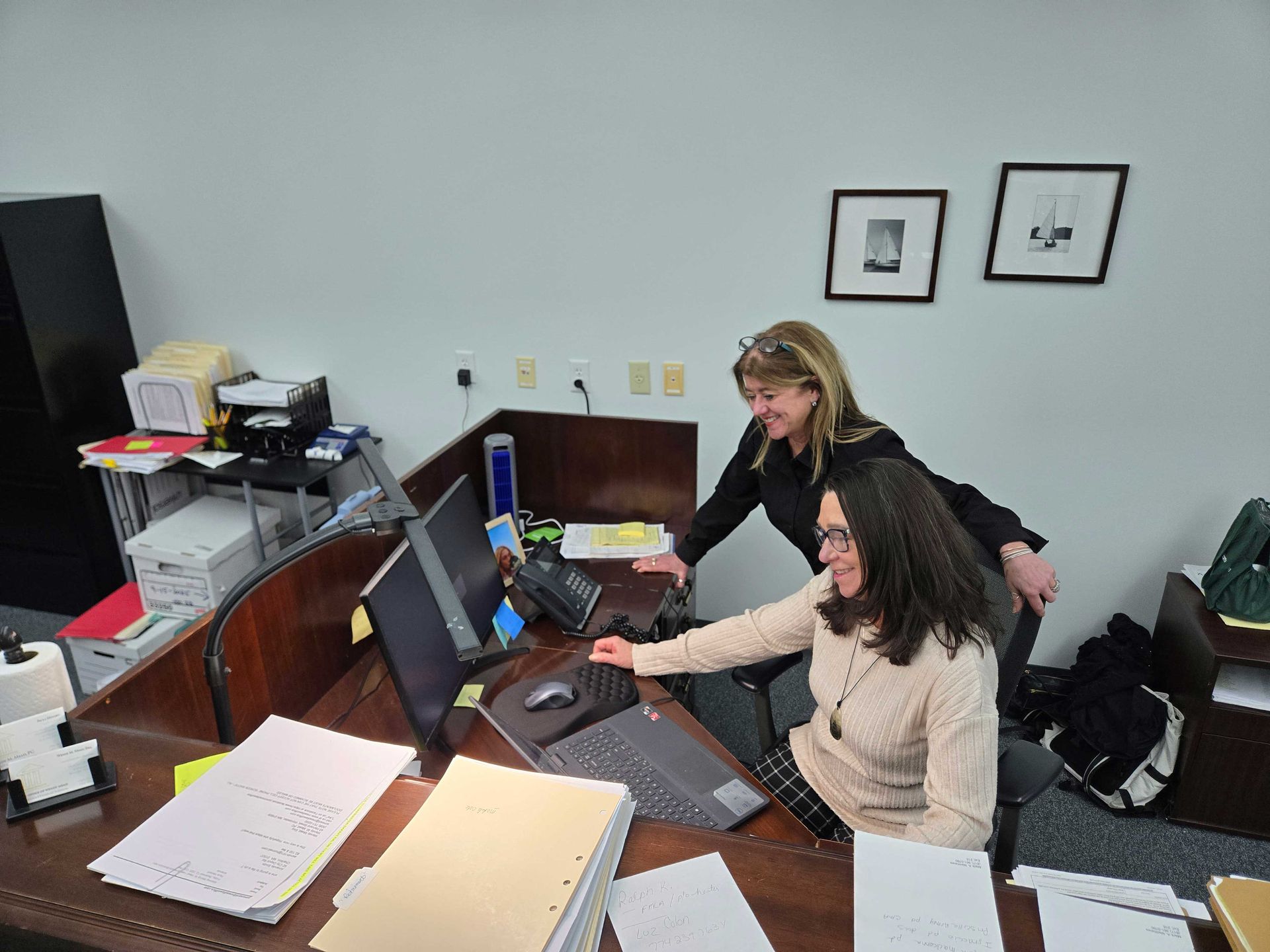 Two people working at a desk with computer, papers, and telephone in an office. One leans over to assist the other.