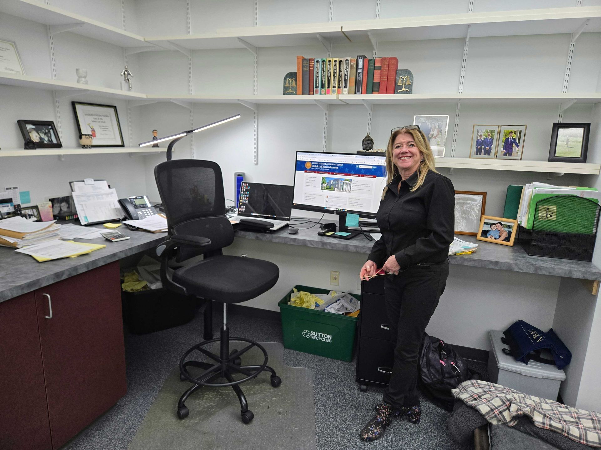 Woman smiles in her office. Bookshelves above a desk with a computer. A desk chair sits in front.