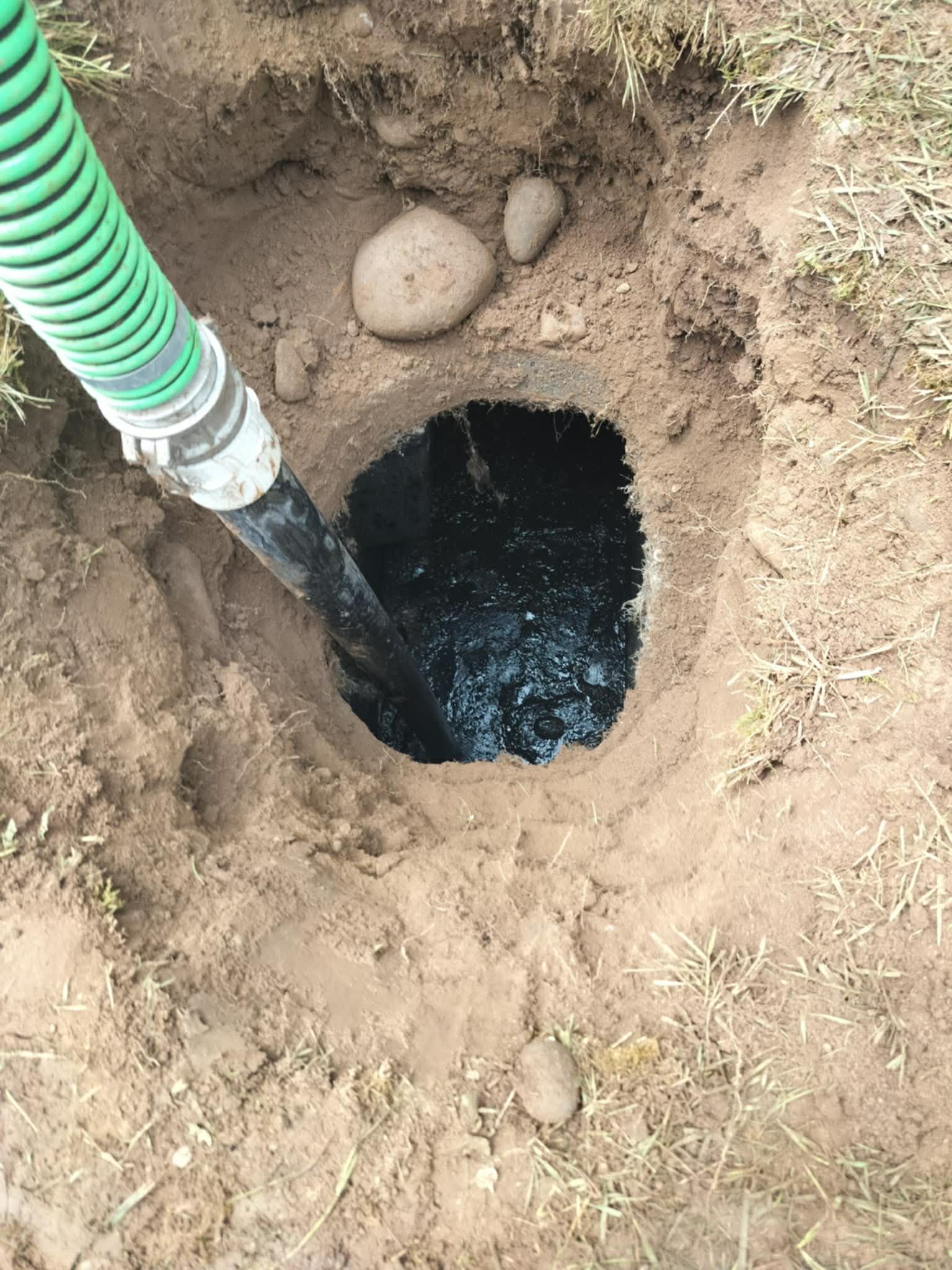 Hole in the ground being pumped out with a green hose, surrounded by dirt and grass.