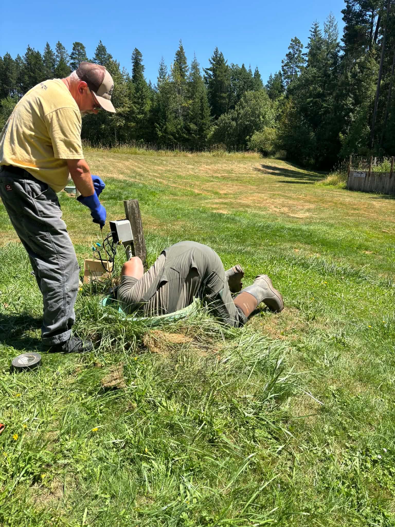 Man working on a ground fixture as another man looks on in a grassy field on a sunny day.