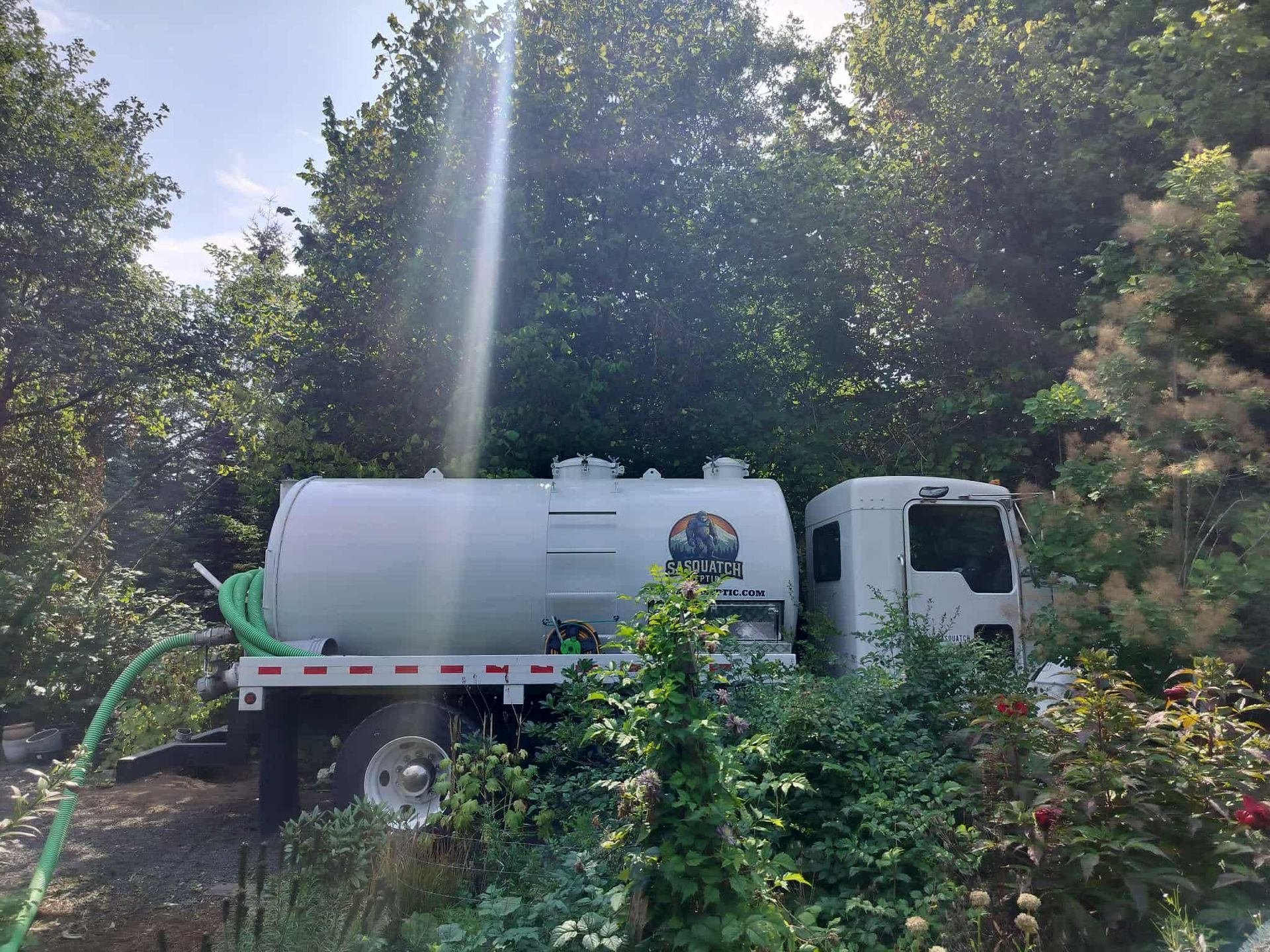 White tank truck parked in a wooded area with trees and sunlight.