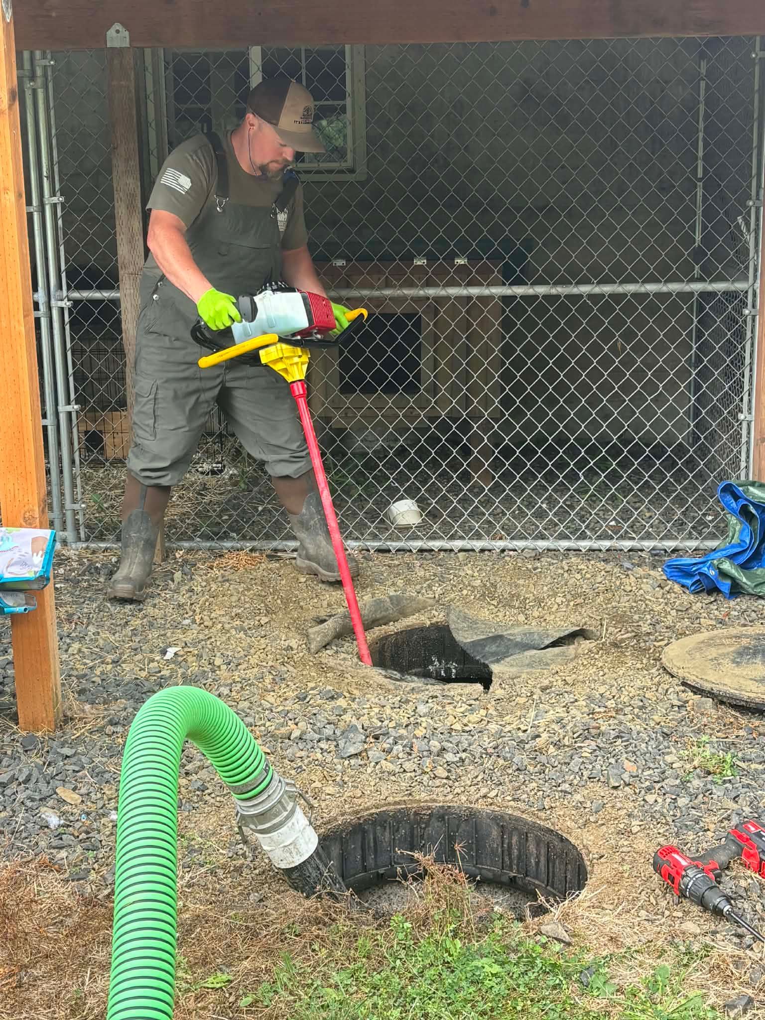 Person using a jackhammer on a tree stump near a chain-link fence. Green hose and safety gear are visible.