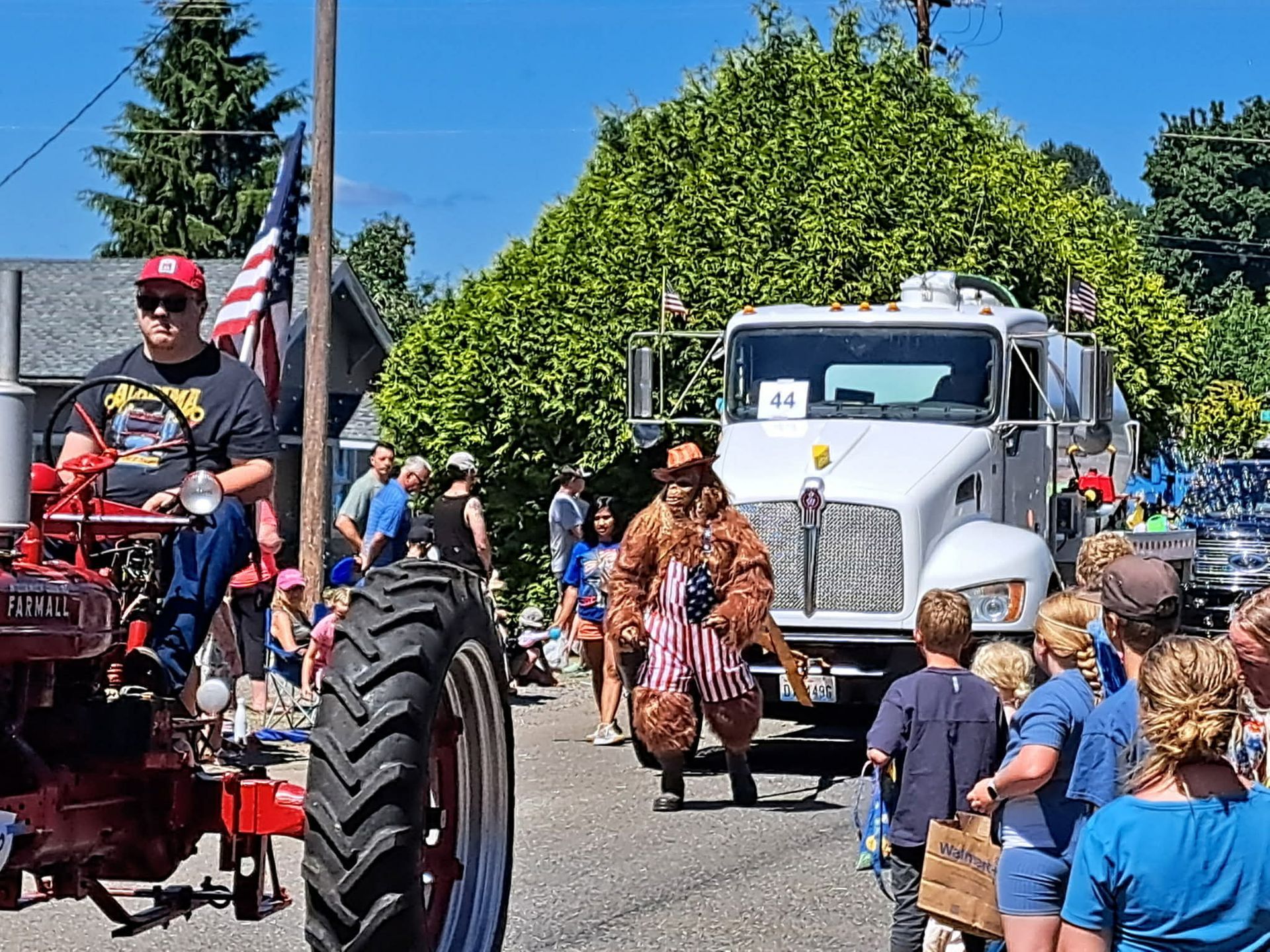 Parade scene: tractor with driver, Bigfoot in American flag vest, white truck, onlookers.