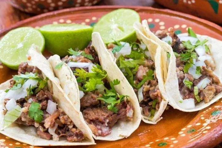 Four tacos filled with meat, topped with chopped onions and cilantro, served on a decorated plate with lime wedges.