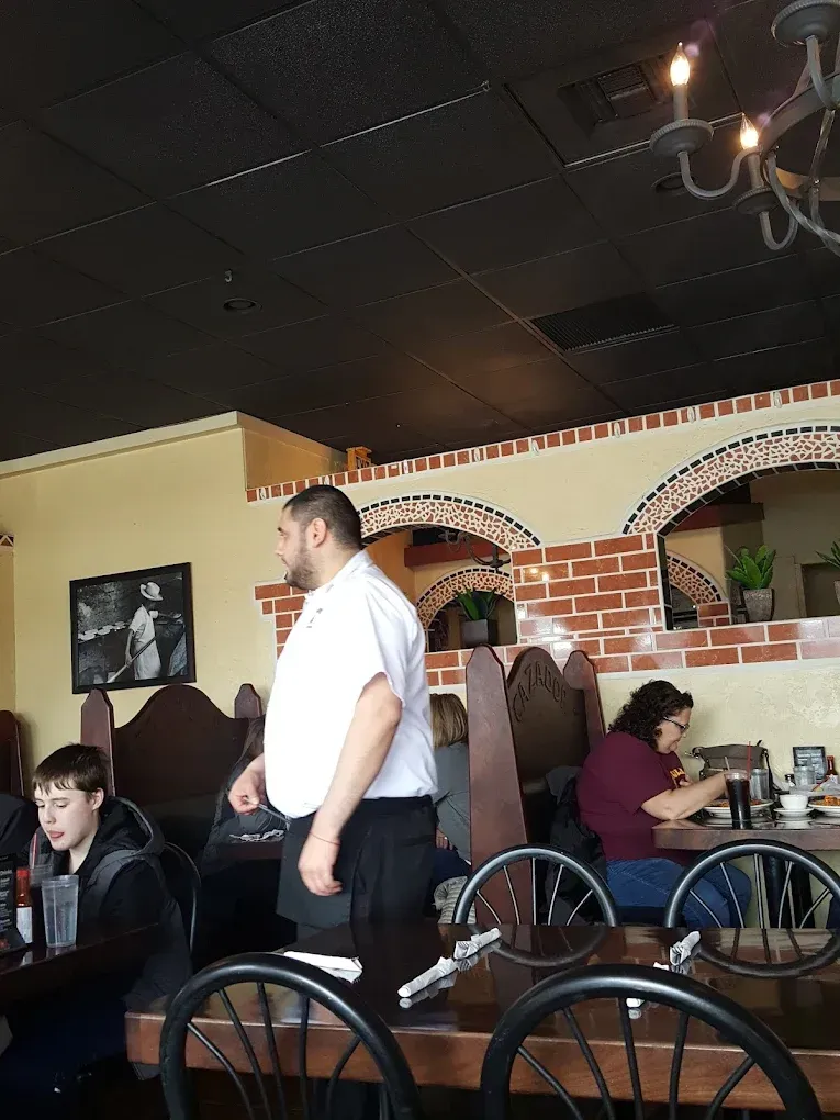 A waiter in a white shirt stands in a restaurant with brick-style decor, near patrons seated at wooden tables.
