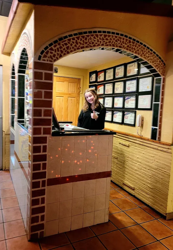 A person smiles behind a tiled reception desk, in front of a wall featuring a grid of framed certificates.