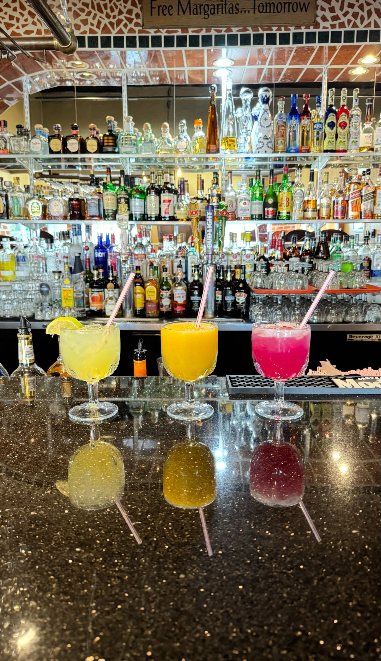 Three colorful margaritas in stemmed glasses lined up on a dark bar counter with a well-stocked liquor shelf behind them.