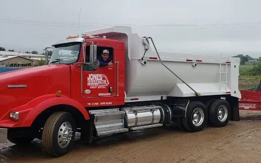 A man is driving a red dump truck on a dirt road.