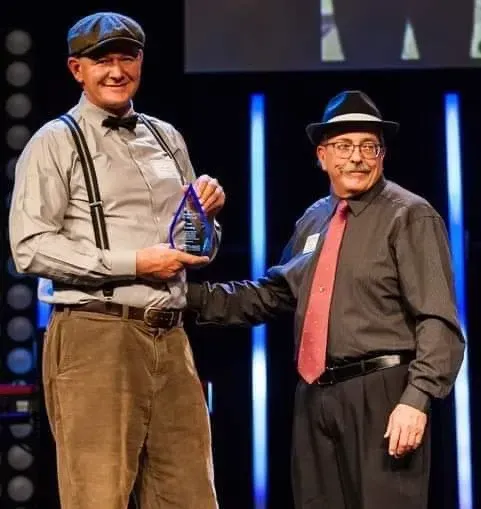 Two men are shaking hands on a stage and one is holding a trophy.
