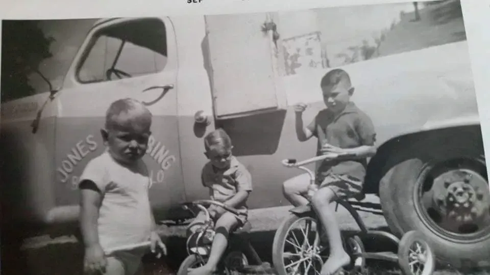 Three children are riding tricycles in front of a jones truck.