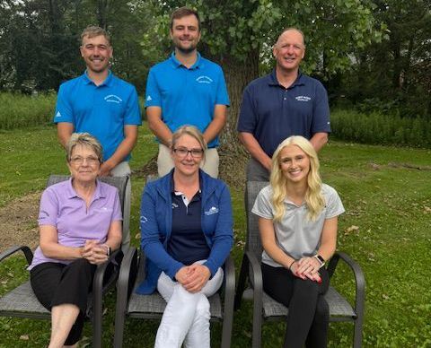 Group of seven people, mostly in blue shirts, posing outdoors. Three seated in chairs, and four standing behind them in front of a tree.