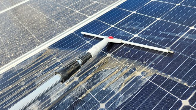 Solar panels being cleaned with a long-handled brush, with water streaks on the surface.