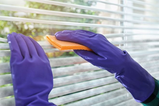 Purple-gloved hands cleaning window blinds with an orange sponge