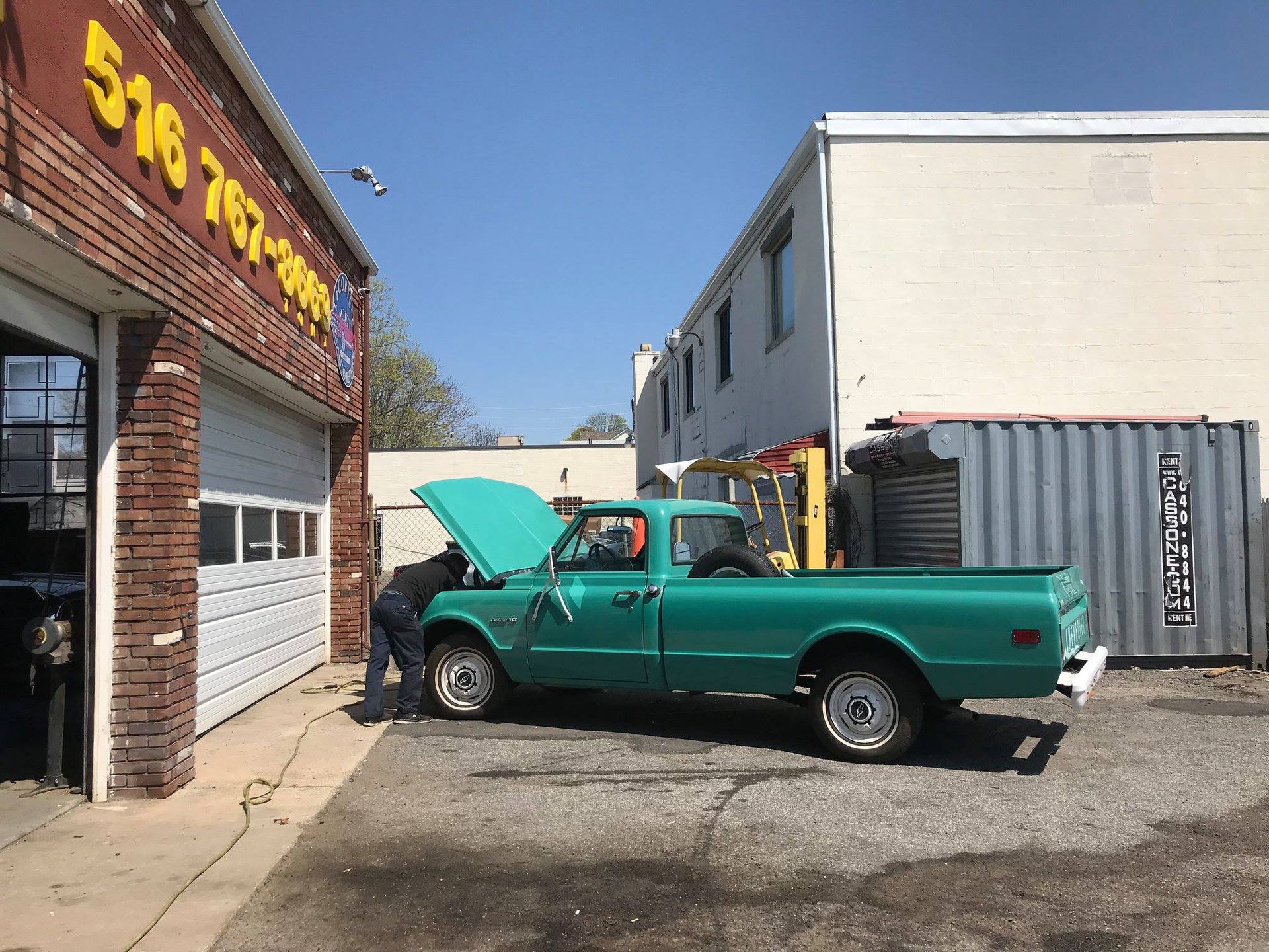 A person working on a teal pickup truck in front of a garage. The truck's hood is open.