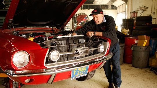 Man working on a red classic Mustang in a garage; hood up.