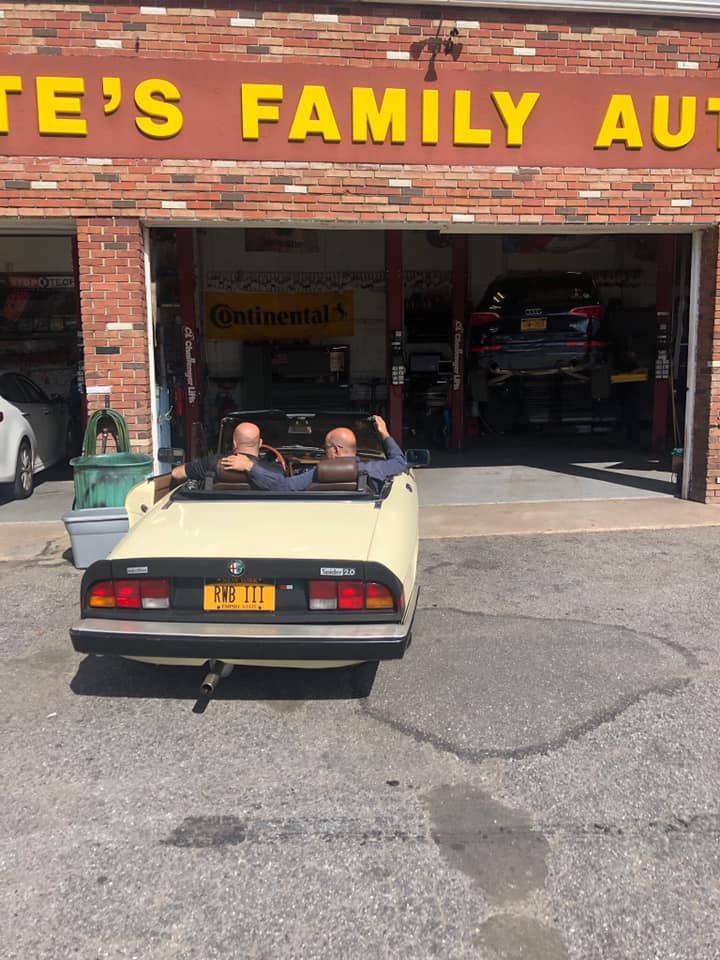 Two men in a cream convertible exit a garage. The sign reads 