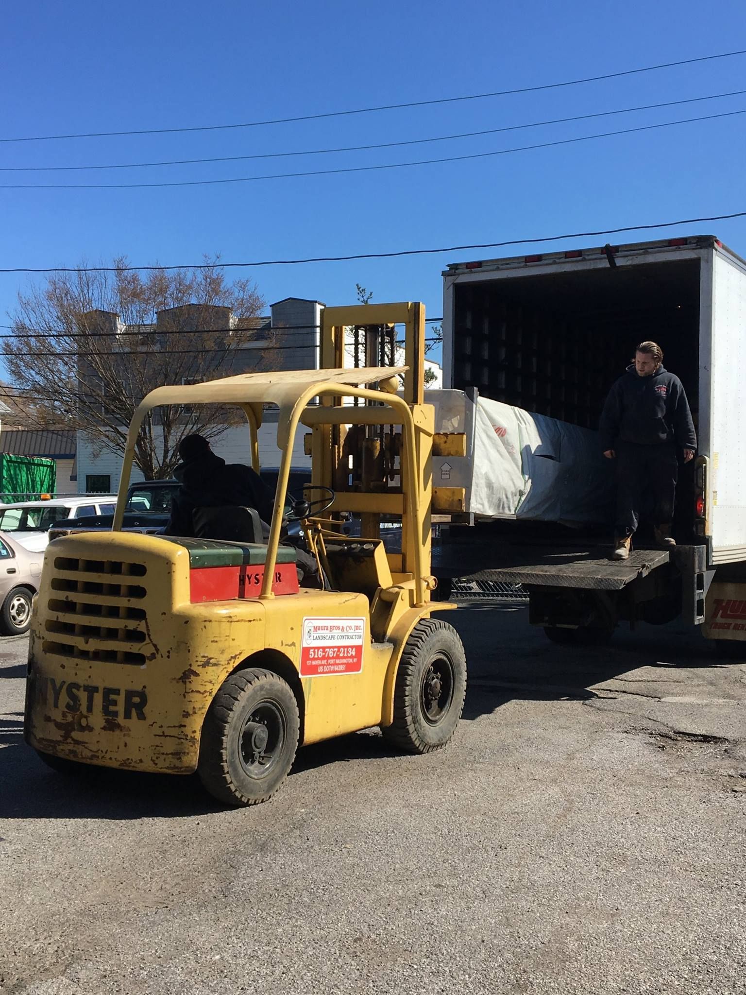 Yellow forklift loading a wrapped item into a white truck. A man stands in the truck. Bright, sunny day.