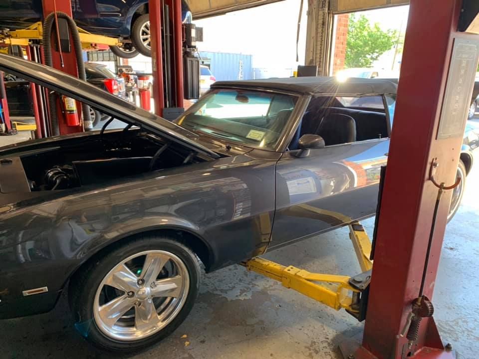 Gray classic car on a lift, hood open in a garage, with a partial view of other cars.