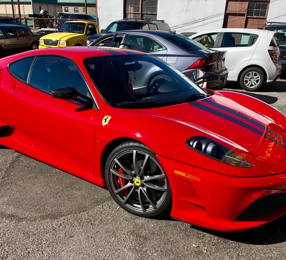 Red Ferrari sports car parked on a paved lot, with black racing stripes and other vehicles in the background.