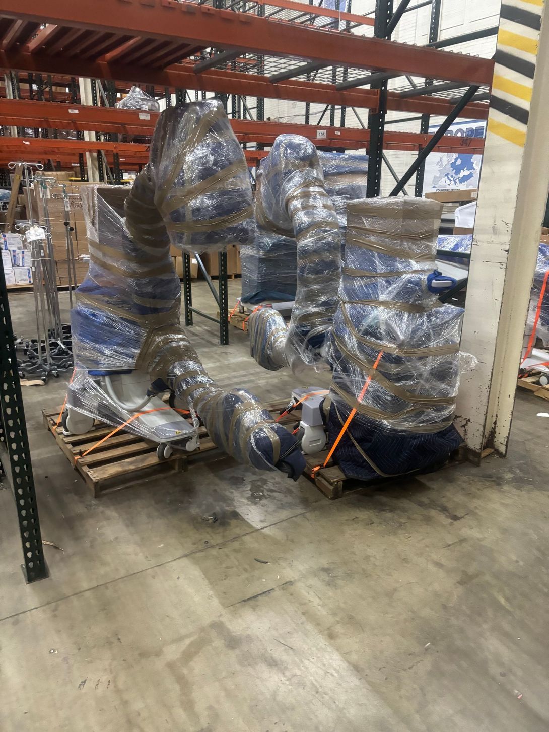 Shrink-wrapped industrial equipment on pallets in a warehouse aisle with orange straps and a striped post.